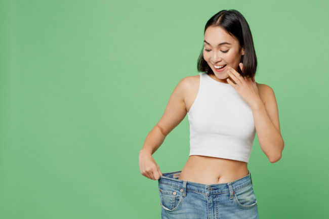 Young happy woman wears white clothes and show loose pants on waist after weight loss in Salt Lake City, UT | Santosh Yoga Institute