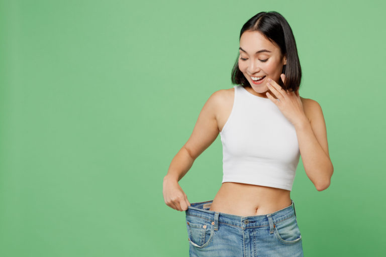 Young happy woman wears white clothes and show loose pants on waist after weight loss in Salt Lake City, UT | Santosh Yoga Institute