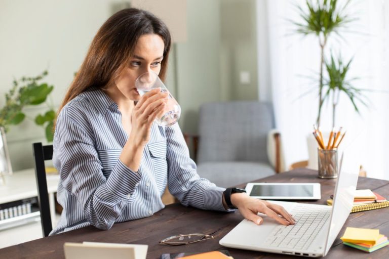 Young woman drinking a glass of water while looking at laptop in Salt Lake City, UT | Santosh Yoga Institute